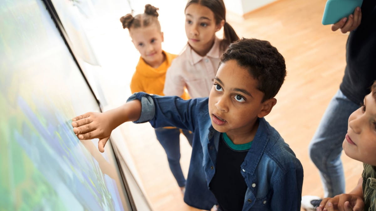 Primary aged students gathered around, and looking up at an interactive display. In the center of the image, a young boy looking amazed at the content on the screen.
