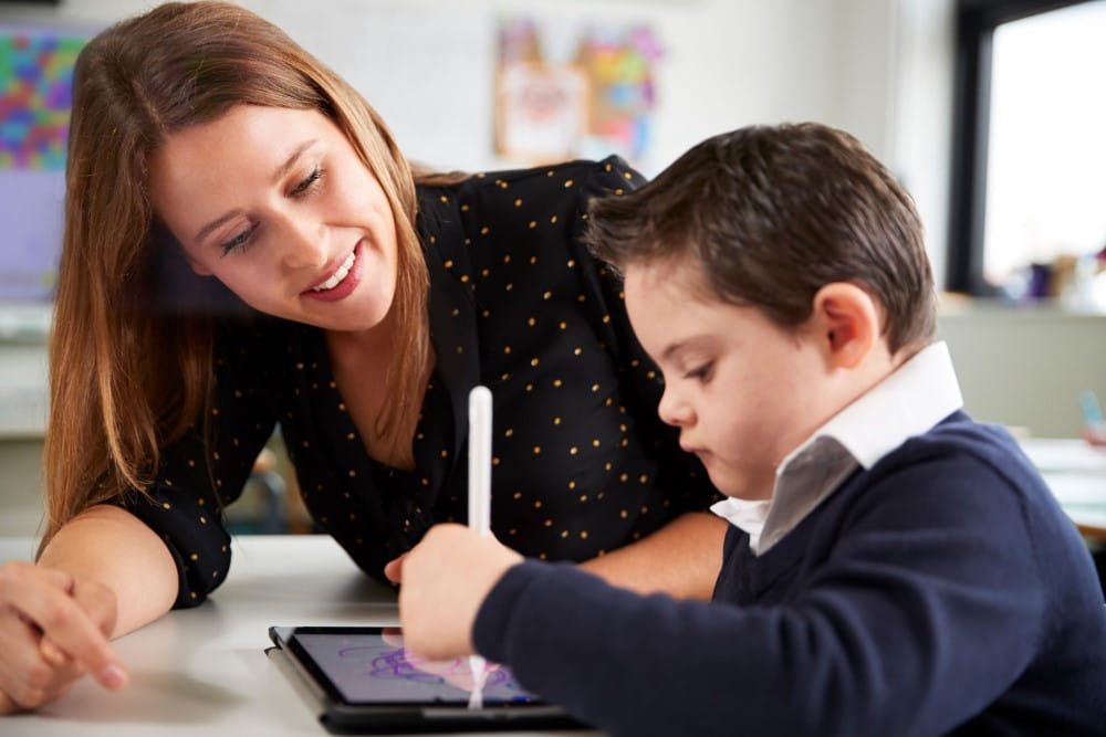 Teacher assisting young student with digital tablet in classroom.