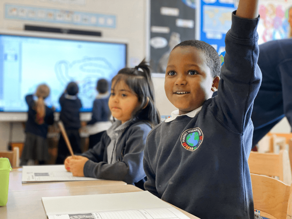 Young student raising hand in the classroom. Studets behind him using an interactive display by SMART.